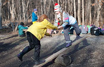 Children playing outdoors - balancing on a plank of wood over a tree stump.