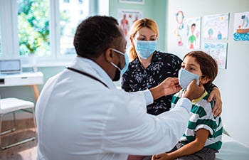A doctor examining a child with the child's mother in the background.